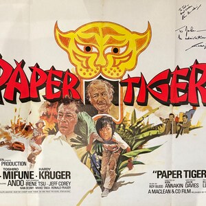 Puede incluir: Un cartel de cine para la película "Paper Tiger" con una cabeza de tigre amarilla con ojos negros y boca roja. El cartel también presenta a los actores David Niven, Toshiro Mifune y Hardy Kruger. El texto "Paper Tiger" está en letras rojas grandes.