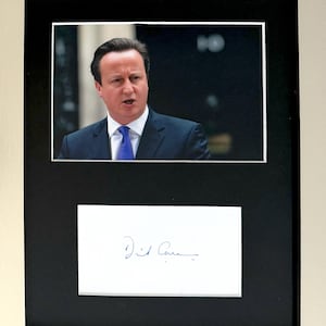 May include: A framed display featuring a photograph of David Cameron wearing a dark suit and blue tie, positioned above a white card with a blue ink signature. The entire arrangement is set within a black mat and a silver-toned frame.