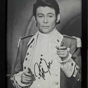 May include: Framed monochrome photograph of a man in a military jacket, holding two small pistols. The jacket features epaulettes and buttons. The photograph is signed in black ink.