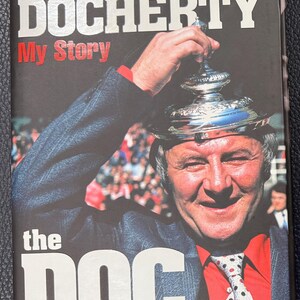 May include: Book cover featuring Tommy Docherty, with the title "Tommy Docherty My Story" in white and red text. The image shows Docherty holding a trophy, wearing a suit and tie. The text "the DOC" and "HALLOWED BE THY GAME" are also visible.