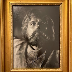 May include: A black and white framed photograph of a man with a beard and long hair. The man is wearing a fringed scarf and has signed his name "Robert Duvall" in the bottom right corner of the photo.