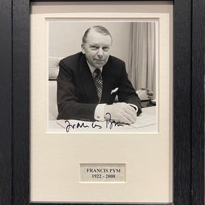 Puede incluir: Fotografía en blanco y negro de un hombre con traje y corbata, sentado en un escritorio. El hombre sonríe y mira a la cámara. La fotografía está firmada "Francis Pym" e incluye las fechas "1922 - 2008".