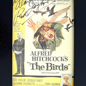 May include: Framed movie poster for Alfred Hitchcock's "The Birds" in a black frame. The poster features a woman screaming, with birds attacking. Includes the film's title, cast, and director's name, with signatures.