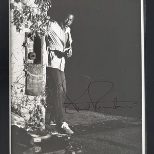May include: Framed black and white photograph of a man in a light jacket and jeans, standing by a wall with climbing plants. The photograph is signed in red ink. The background is dark.