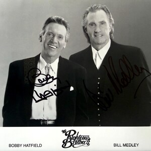 May include: Black and white photograph of The Righteous Brothers, Bobby Hatfield and Bill Medley, in suits. Both men are smiling, and their autographs are visible. The Righteous Brothers logo and names are at the bottom.