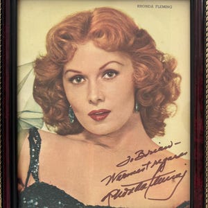 Może przedstawiać: Oprawiony portret Rhondy Fleming z kasztanowymi włosami, noszącej ciemne kolczyki i ciemny top z cekinami. Nadruk zawiera imię i nazwisko Rhonda Fleming u góry oraz odręczny podpis ciemnym tuszem w dolnej części obrazu.