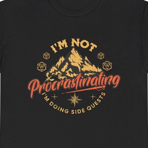 May include: Black t-shirt with a yellow and orange graphic of a mountain range with dice and the text "I'm not procrastinating I'm doing side quests."