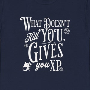 Puede incluir: Camiseta azul marino con texto blanco que dice "What Doesn't Kill You, Gives You XP". El texto está en una fuente estilizada e incluye un gráfico de dragón y dados.