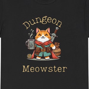 Può includere: Maglietta nera con un gatto dei cartoni animati che indossa un mantello rosso e tiene una tazza e una spada. Il testo "Dungeon Meowster" è stampato sotto il gatto.