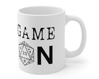 May include: White ceramic coffee mug with black text that reads "GAME ON" and a black and white illustration of a twenty-sided die.