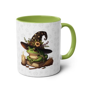 May include: A green frog wearing a witch's hat with a floral design and holding a book. The frog is sitting on a book with a candle and a broom next to it. The mug has a green rim and a white background with a pattern of mushrooms.