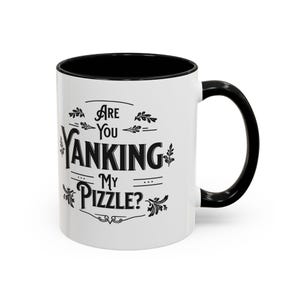 Könnte beinhalten: Weiße Keramik-Kaffeetasse mit schwarzem Innenraum und Henkel. Die Tasse hat ein schwarzes Motiv mit der Aufschrift "Are you yanking my pizzle?" mit dekorativen Verzierungen.