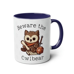 Peut inclure: Une tasse en céramique blanche avec un bord et une poignée bleus. La tasse représente un hibou de dessin animé avec une épée et un dé, et le texte "Beware the Owlbear".