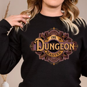 Puede incluir: Sudadera negra con un diseño gráfico morado y dorado que dice "The Dungeon Master". El gráfico presenta una espada estilizada, dados y otros elementos de fantasía.