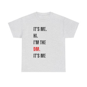 Peut inclure: Un t-shirt gris avec le texte "IT'S ME. HI. I'M THE DM. IT'S ME" imprimé dessus. Le mot "DM" est en rouge.