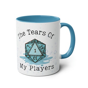 Op de afbeelding: Witte keramische mok met een blauw interieur en handvat. De mok heeft de tekst "The Tears Of My Players" en een gestileerde D20-dobbelsteenafbeelding in de kleur teal. Waterdruppelafbeeldingen zijn rond de mok verspreid.