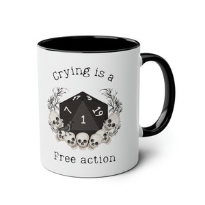 Op de afbeelding: Een witte keramische mok met een zwarte rand en handvat. De mok heeft een zwart-witte illustratie van een 20-zijdige dobbelsteen omringd door schedels en een raaf. De tekst "Crying is a Free action" staat op de mok gedrukt.