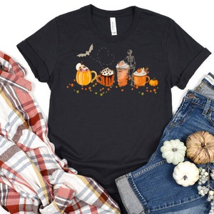 May include: Black t-shirt featuring a graphic of four autumn-themed coffee mugs with a skeleton holding a mug and a bat flying above. The mugs are decorated with pumpkins, leaves, and whipped cream.