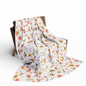 May include: A white throw blanket featuring a watercolour pattern of mushrooms, leaves, and flowers in shades of orange, yellow, and brown. The blanket is draped over a brown armchair.