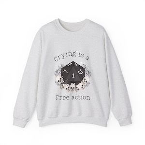 Op de afbeelding: Een grijs sweatshirt met de tekst "Crying is a free action" erop gedrukt. Een zwart-witte illustratie van een 20-zijdige dobbelsteen met schedels en een raaf is gecentreerd op het sweatshirt.