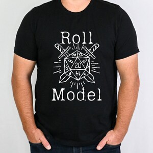 May include: Black t-shirt featuring a white graphic of a 20-sided die with crossed swords and the text "Roll Model".