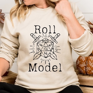 May include: A cream-colored sweatshirt with a black graphic of a twenty-sided die with two swords crossed behind it. The text "Roll Model" is printed below the die.