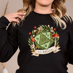 Könnte beinhalten: Schwarzes Sweatshirt mit einem grünen 20-seitigen Würfel, umgeben von Blumen und Pilzen. Der Würfel hat die Zahlen 2, 12, 20, 6, 8 und 16. Der Text "All Natural" befindet sich auf einem weißen Banner.