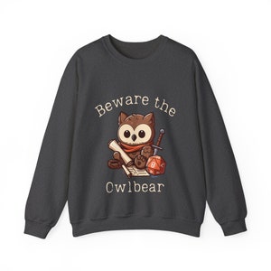 Könnte beinhalten: Ein dunkelgraues Sweatshirt mit dem Text "Beware the Owlbear" auf der Vorderseite. Der Text ist weiß und von einem Cartoon-Bild eines Eulenbären umgeben, der ein Schwert und eine Schriftrolle hält.