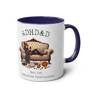 Könnte beinhalten: Weiße Keramiktasse mit blauem Rand. Die Tasse zeigt eine Comic-Illustration einer Person, die auf einem Sofa sitzt, mit Würfeln und einem Zauberstab, die um sie herum verstreut sind. Der Text "ADHD & D" befindet sich oben auf der Tasse und "Roll for Executive Dysfunction" unten.