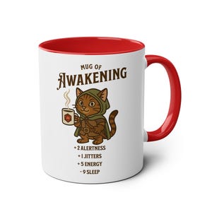 May include: White ceramic mug with a red rim and handle. The mug features a cartoon cat wearing a green hooded cloak and holding a mug with a 20-sided die. The text "MUG OF AWAKENING" is above the cat and the text "+ 2 ALERTNESS + 1 JITTERS + 5 ENERGY - 9 SLEEP" is below the cat.