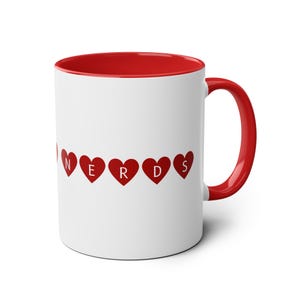 Könnte beinhalten: Weiße Keramiktasse mit rotem Rand und Henkel. Die Tasse hat das Wort "NERDS" in roten Herzen geschrieben.