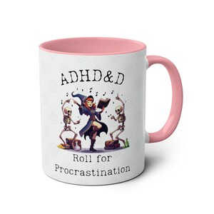 May include: White ceramic mug with a pink rim and handle. The mug features a cartoon illustration of a witch with two skeletons dancing. The text on the mug reads "ADHD&D Roll for Procrastination".