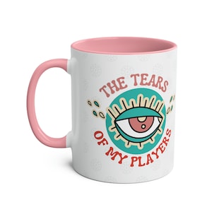 May include: White ceramic coffee mug with a pink rim and handle. The mug features a teal and red eye with the text "The Tears of My Players" around it.