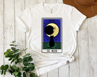 Puede incluir: Camiseta blanca con un gato negro mirando a una luna amarilla con cara. La imagen es una carta del tarot con el texto "XVIII THE MOON".