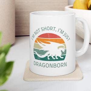 May include: White ceramic mug with a handle, featuring a retro design of a dragon silhouette inside a colorful circle. The text reads "I'M NOT SHORT, I'M JUST DRAGONBORN" in green and white.