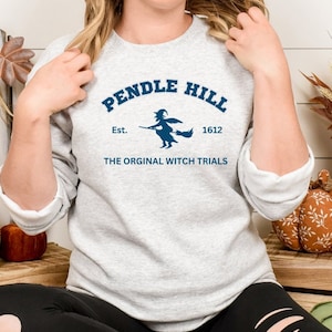 May include: A gray sweatshirt with a blue graphic print of a witch flying on a broomstick. The graphic includes the text "Pendle Hill Est. 1612 The Original Witch Trials".