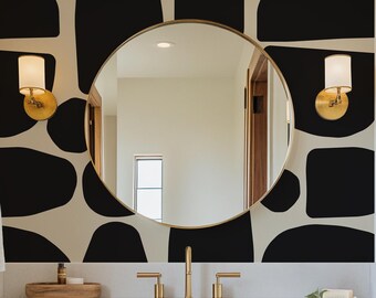 May include: A modern bathroom vanity with a white countertop and wooden cabinets. The vanity features a round mirror with a gold frame, two gold faucets, and a black and white geometric wallpaper.
