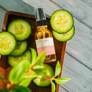 Peut inclure: Un flacon en verre transparent avec un compte-gouttes rempli d'un liquide jaune. Le flacon est étiqueté "Skin Supplie" et "Cucumber Face Serum". Le flacon est posé sur un plateau en bois avec des tranches de concombre.