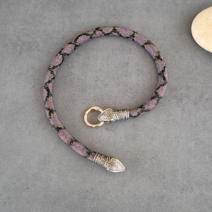 Puede incluir: Un collar de cuentas flexible con un patrón de diamantes en violeta y negro. Los extremos terminan en cabezas de serpiente de metal plateado, una de las cuales sujeta un cierre circular dorado que se conecta a la otra cabeza para cerrar el aro.