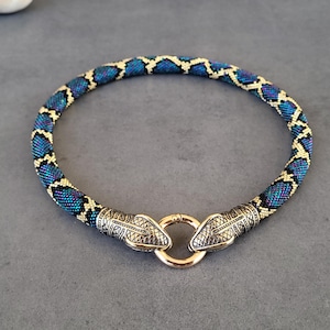 May include: A beaded necklace with a snake scale pattern in blue, teal, and gold. The necklace has a gold clasp and two silver snake head accents. The jewelry is designed to be worn around the neck.
