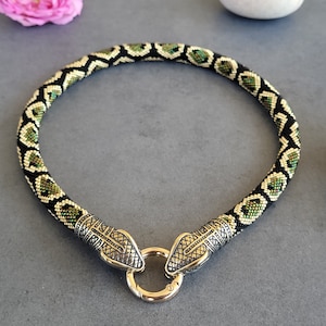 May include: A beaded necklace with a snake design. The necklace has a black, green, and gold beaded pattern. The ends of the necklace are detailed with silver-coloured snake heads and a gold-coloured clasp. The necklace is displayed on a grey surface.