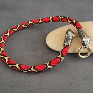Peut inclure: Un collier de perles avec un motif de serpent. Le collier présente un motif rouge, noir et or. Le fermoir est de couleur argentée et en forme de tête de serpent. Le collier mesure environ 40 cm de long.