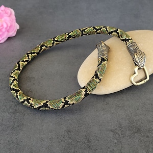 Könnte beinhalten: Eine Halskette aus Perlen mit einem schlangenartigen Muster in Grün, Schwarz und Gold. Die Halskette hat Metall-Schlangenkopf-Terminals und einen herzförmigen Verschluss. Eine rosa Rose und ein glatter Stein im Hintergrund.