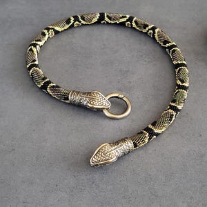Pode incluir: Um colar flexível de missangas com um padrão repetitivo de pele de cobra em preto e dourado. As extremidades são rematadas com terminais metálicos detalhados em forma de cabeça de cobra em tom de latão, com um anel de fecho circular.