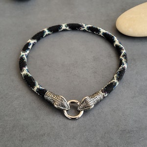 Puede incluir: Un collar de cuentas con diseño de serpiente. El collar presenta un patrón en blanco y negro con cierres de cabeza de serpiente plateados y un cierre de anillo plateado. El collar mide aproximadamente 40 cm de largo.
