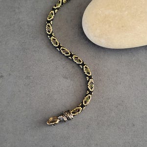 May include: A flexible beaded bracelet shaped like a snake, featuring a repeating pattern of black, gold, and olive green seed beads. The bracelet includes a metallic snake head at one end and a metallic hook clasp at the other end.