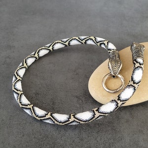 Peut inclure: Un collier de perles avec un motif de serpent. Le collier présente un motif de perles blanches, noires et dorées. Les extrémités du collier sont ornées de fermoirs en forme de tête de serpent argentés et d'un fermoir en forme d'anneau argenté.