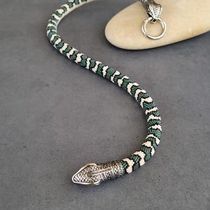 Könnte beinhalten: Eine flexible, schlangenförmige Halskette oder Kette mit einem perlenbesetzten Körper in einem sich wiederholenden Muster aus dunkelgrünen, hellgrünen und cremefarbenen Perlen. Das Stück hat zwei detaillierte silberfarbene Schlangenköpfe an den Enden, wobei ein Kopf an einem runden Ringverschluss befestigt ist.