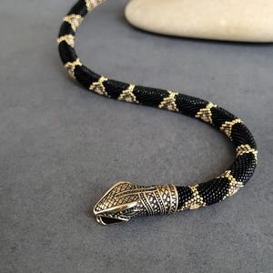 Może przedstawiać: Naszyjnik w kształcie węża z czarnych i złotych koralików. Ciało węża jest czarne ze złotymi wzorami w kształcie rombów. Głowa jest ze złotego metalu z misternymi wzorami. Na szarym tle.