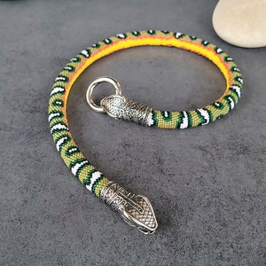 Peut inclure: Un bracelet en perles flexible au motif de serpent avec des couleurs jaune, vert, blanc et noir. Le bijou est terminé par deux embouts en métal argenté en forme de tête de serpent, dont l'un est doté d'un fermoir à anneau circulaire.